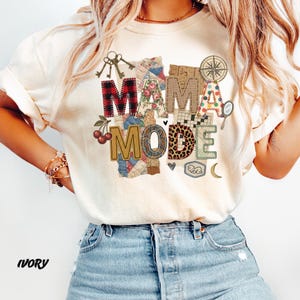 May include: Ivory-colored t-shirt with the words "MAMA MODE" in a patchwork design. The letter "M" is red plaid, and the letter "A" is floral. The shirt has a crew neck and short sleeves.