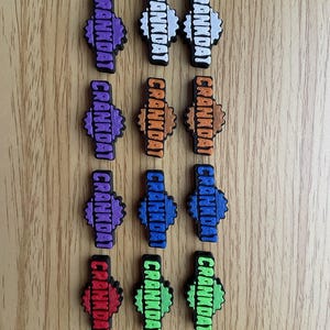 May include: A collection of colorful shoe charms with the word "CRANKDAT" in white block letters. The charms are in various colors, including purple, white, orange, blue, red, and green. The charms are arranged on a light brown wooden surface.