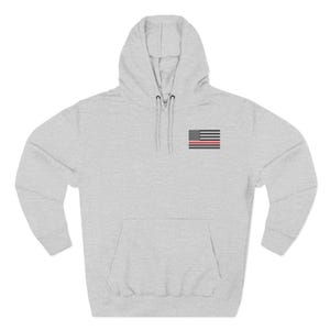 May include: A light gray hoodie with a front pocket and a hood. The hoodie features a small American flag patch with a red stripe, symbolizing support for firefighters. The design is simple and suitable for casual wear.