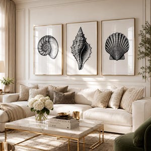 May include: Three framed wall art prints featuring detailed black and white illustrations of seashells. Each print is encased in a thin gold frame and displayed in a horizontal row on a light-colored wall above a large beige sectional sofa.