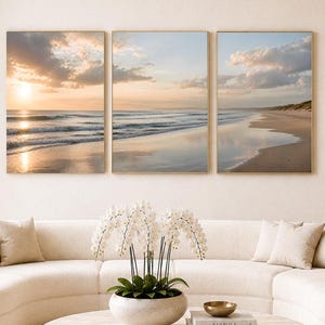 以下が含まれることがあります： 夕暮れのビーチ、海の波、砂浜を描いた3枚組のウォールアートセット。クリーム色の曲線的なソファ、白い蘭が飾られた丸い石のコーヒーテーブル、そしてお揃いの肘掛け椅子が配置された明るい部屋の壁に飾られています。
