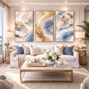 May include: Three framed abstract art pieces with gold frames. The art features watercolor designs in shades of blue, beige, and gold. A white sofa with blue and beige pillows sits below the artwork. A coffee table and side tables complete the living room setting.