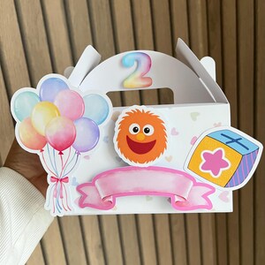 Puede incluir: Caja de regalo blanca decorada con pegatinas coloridas, incluyendo globos, un personaje naranja difuso y un bloque en forma de estrella. El n&uacute;mero "2" se muestra en la parte superior. Tambi&eacute;n se incluye una pancarta rosa.