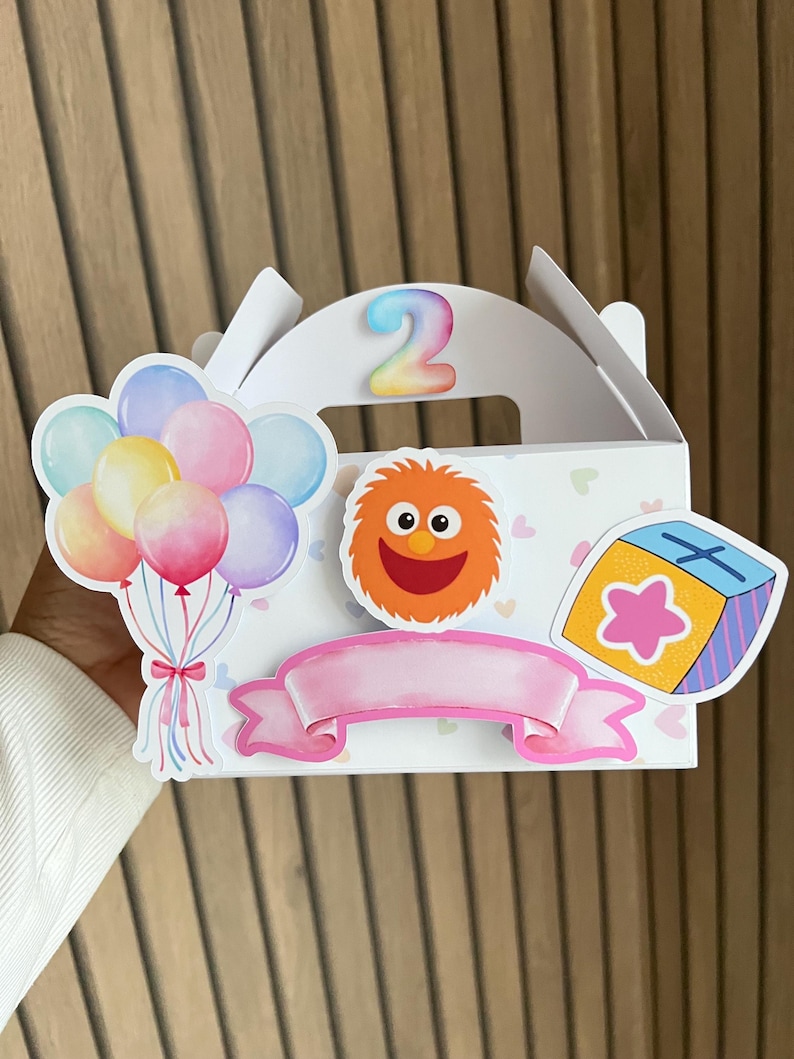 Puede incluir: Caja de regalo blanca decorada con pegatinas coloridas, incluyendo globos, un personaje naranja difuso y un bloque en forma de estrella. El n&uacute;mero "2" se muestra en la parte superior. Tambi&eacute;n se incluye una pancarta rosa.