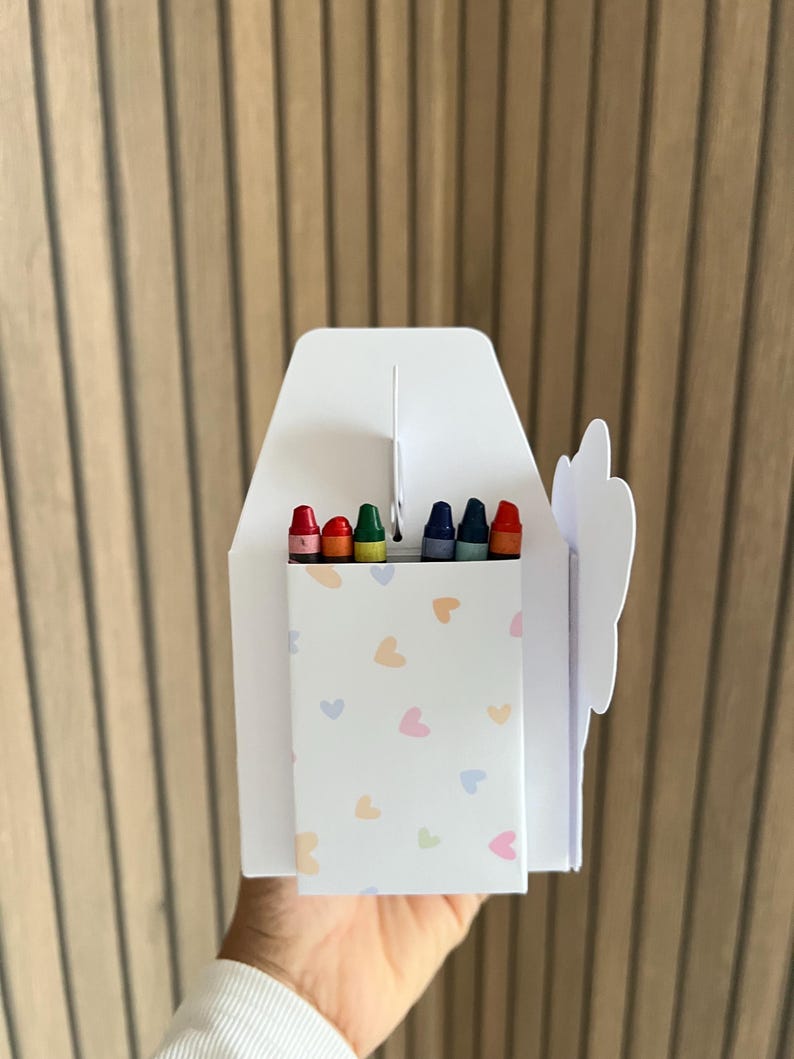 Puede incluir: Una caja blanca de crayones con cierre de solapa, que contiene seis crayones de colores. La caja est&aacute; decorada con corazones pastel y tiene un recorte en forma de nube. Los crayones son rojos, naranja, verdes y azules.
