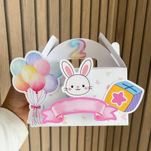 Puede incluir: Una caja de regalo blanca con un conejito, globos y una pancarta rosa. El n&uacute;mero "2" est&aacute; en la parte superior. La caja tiene un asa y est&aacute; decorada con ilustraciones coloridas, ideal para un cumplea&ntilde;os infantil.