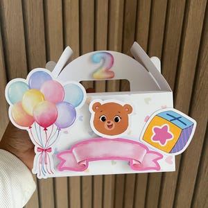 Puede incluir: Una caja de regalo blanca con asa, decorada con un oso de dibujos animados, globos y una pancarta rosa. El n&uacute;mero "2" se muestra en la parte superior. La caja tiene un dise&ntilde;o colorido con tem&aacute;tica de cumplea&ntilde;os.