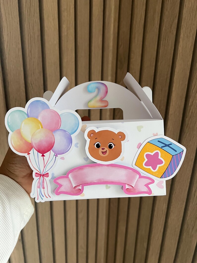 Puede incluir: Una caja de regalo blanca con asa, decorada con un oso de dibujos animados, globos y una pancarta rosa. El n&uacute;mero "2" se muestra en la parte superior. La caja tiene un dise&ntilde;o colorido con tem&aacute;tica de cumplea&ntilde;os.