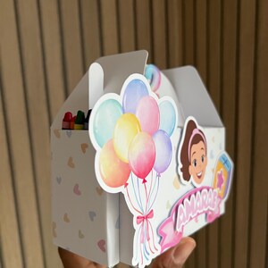 Puede incluir: Una caja de recuerdos blanca con asa, decorada con globos de colores y un personaje de dibujos animados. La caja tiene un patr&oacute;n de corazones y el nombre "AMARIE" en una pancarta rosa. Se ven crayones dentro.