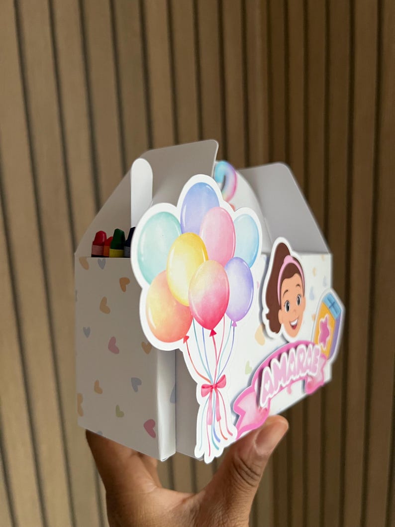 Puede incluir: Una caja de recuerdos blanca con asa, decorada con globos de colores y un personaje de dibujos animados. La caja tiene un patr&oacute;n de corazones y el nombre "AMARIE" en una pancarta rosa. Se ven crayones dentro.