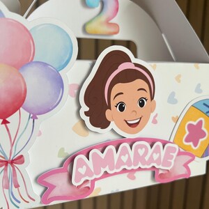 Puede incluir: Una caja de regalo blanca decorada con una ni&ntilde;a de dibujos animados, globos y el nombre "AMARAE" en letras rosas. Un n&uacute;mero colorido "2" est&aacute; en la parte superior, lo que sugiere una celebraci&oacute;n de segundo cumplea&ntilde;os.