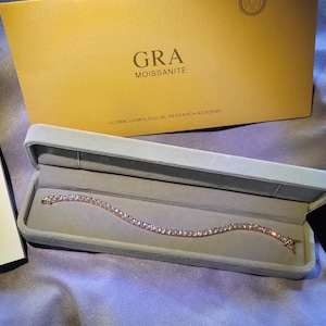 以下が含まれることがあります： 複数の宝石が付いた輝くブレスレットが、開いた薄いグレーのジュエリーボックスに展示されています。ボックスは紫色の表面にあり、GRAモアッサナイトレポートとGRAモアッサナイトカードの隣にあります。「GRA Moissanite」と書かれた黄色のボックスが背景にあります。