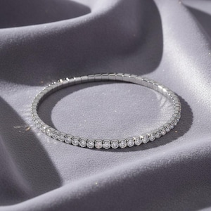 Puede incluir: Una pulsera plateada con una fila de pequeñas piedras preciosas transparentes. La pulsera se presenta sobre un fondo gris sedoso, creando una estética lujosa y elegante. Las piedras preciosas brillan.