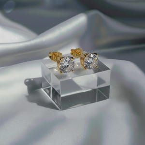 May include: A pair of gold-colored stud earrings with large, round, clear gemstones. The earrings are displayed on a clear, square block against a backdrop of draped, white fabric. The earrings are set in a gold-colored metal.