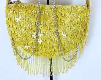 May include: Yellow beaded shoulder bag with a floral design. The bag features a beaded fringe and a silver chain strap. The bag is decorated with yellow beads and small white flower accents.