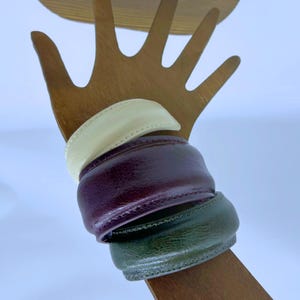 Può includere: Tre braccialetti in pelle color crema, bordeaux e verde oliva sono esposti su un porta gioielli a forma di mano in legno. I braccialetti hanno un design arrotondato e sono impilati l'uno sull'altro.
