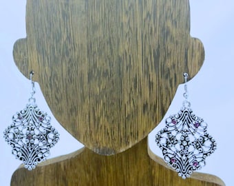 Op de afbeelding: Zilverkleurige oorhangers met een sierlijk, opengewerkt ontwerp. Elke oorbel heeft een ruitvormig frame met kleine, ronde, heldere en roze edelstenen. De oorbellen worden getoond op een houten sieradenstandaard.