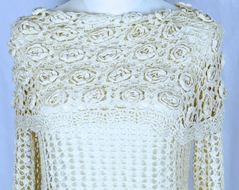 May include: Cream-colored crochet top with long sleeves. The top features a detailed floral pattern around the neckline and shoulders. The open weave design of the sleeves and body creates a delicate, airy look. The garment is displayed on a mannequin.
