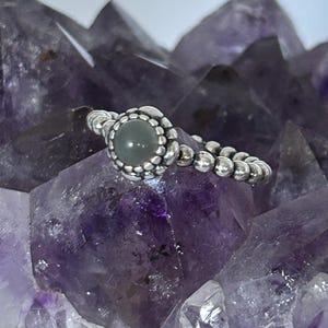 May include: A silver ring with a round, pale green stone set in a beaded bezel. The ring sits on a purple amethyst crystal, showcasing its intricate design and color contrast. The ring's band is also made of small silver beads.
