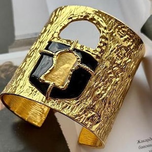 May include: A wide, gold-toned cuff bracelet with a textured surface. The bracelet features a black and gold enamel design in the center, with a cutout detail. The cuff is open at the bottom, allowing it to be slipped onto the wrist.