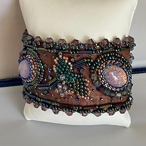 May include: A wide cuff bracelet featuring a brown cork base adorned with iridescent seed beads, dark metallic tube beads, and two large oval cabochons. One cabochon is a pale pink stone, while the other displays a shimmering, multi-colored opal effect.