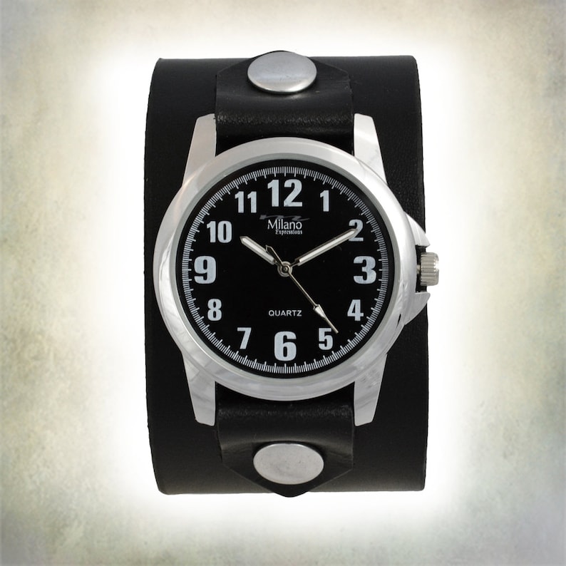 K&ouml;nnte beinhalten: Eine schwarze Lederarmbanduhr mit silbernem Zifferblatt und schwarzen Zeigern. Das Zifferblatt der Uhr hat wei&szlig;e Zahlen und die Worte "Milano Experience" und "QUARTZ".