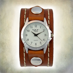 Puede incluir: Un reloj de pulsera con brazalete de cuero marr&oacute;n y esfera plateada. La esfera tiene agujas negras y el texto "Milano Expressions Quartz". El brazalete se asegura con dos tachuelas plateadas y costuras blancas.