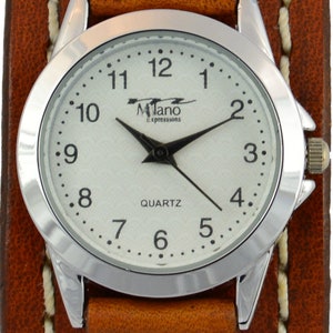 Puede incluir: Un reloj de pulsera con brazalete de cuero marr&oacute;n con esfera plateada. La esfera tiene agujas negras y el texto "Milano Expressions QUARTZ". La correa ancha tiene tachuelas plateadas.