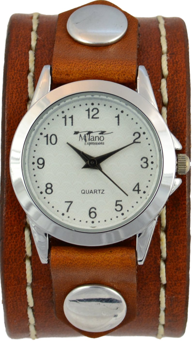 Puede incluir: Un reloj de pulsera con brazalete de cuero marr&oacute;n con esfera plateada. La esfera tiene agujas negras y el texto "Milano Expressions QUARTZ". La correa ancha tiene tachuelas plateadas.