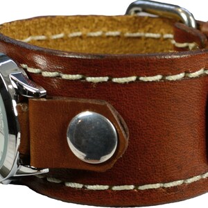 Puede incluir: Un reloj de pulsera con brazalete de cuero marr&oacute;n con esfera plateada. La correa del reloj es de cuero grueso cosido con interior marr&oacute;n claro. La esfera tiene n&uacute;meros y agujas negros. La correa se asegura con una hebilla plateada y un broche plateado.