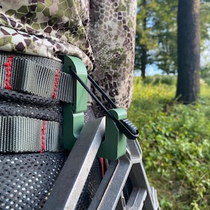 Puede incluir: Primer plano de equipo de caza, con una ayuda para escalar de metal verde sujeta a una mochila de camuflaje. La ayuda está conectada con un cordón elástico negro. La mochila tiene correas grises con costuras rojas y una parte trasera de malla negra.