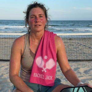 May include: A woman wearing a gray tank top and sitting on a sandy beach with a pink towel draped over her shoulder. The towel features a pickleball design and the name "RACHEL JACK". A pickleball paddle is visible.