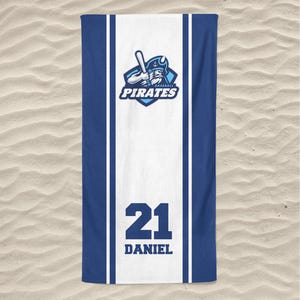 May include: A blue and white beach towel featuring the text "PIRATES" and a baseball bat logo. The towel has a white stripe down the center with the number "21" and the name "DANIEL" printed in black.