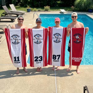 May include: Four people holding red and white baseball towels. Each towel has the text "Baseball Maniacs" and a baseball player graphic. The towels display numbers 11, 23, and 87, and the name "Coach Max".