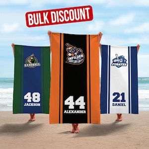 May include: Three beach towels with baseball-themed designs are held up on a beach. The towels feature team logos, player numbers, and names. One towel is green with a baseball logo, another is orange and black with an Infielder logo, and the third is white and blue with a Pirates logo. The text "BULK DISCOUNT" is in a red banner.