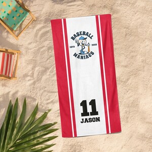 May include: A red and white baseball towel with the text "Baseball Maniacs" and the number "11" with the name "Jason". The towel has a cartoon baseball player logo and is on a sandy surface.