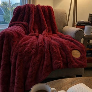 May include: A deep red faux fur throw blanket with a circular tan leather patch featuring the text M-Rose Home & Living. The blanket has a thick, textured pile and is draped over a neutral-colored armchair in a living room setting.