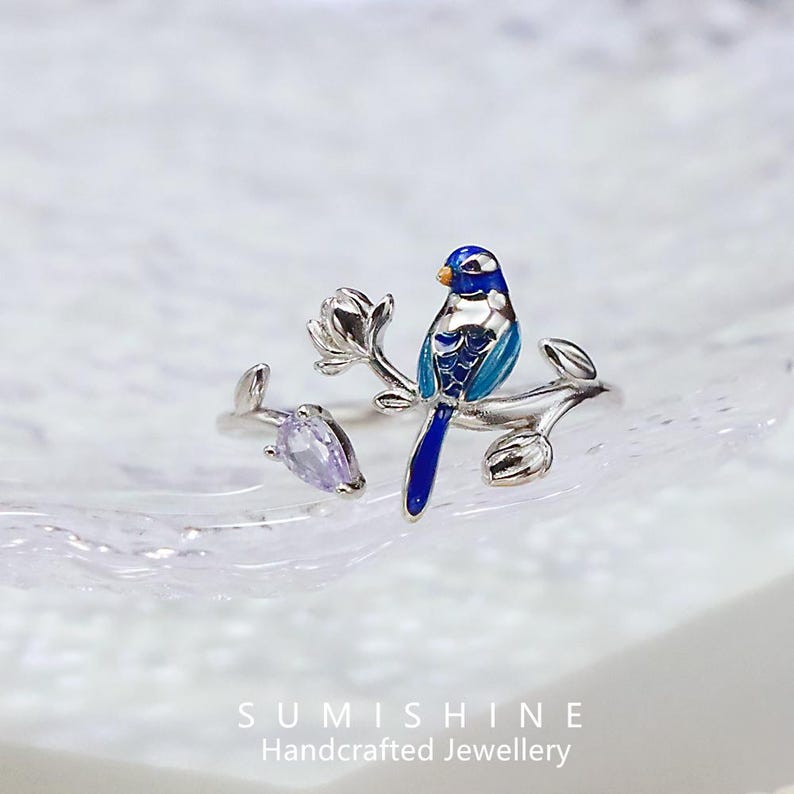 May include: A silver ring featuring a blue and white bird perched on a branch with a flower and a purple gemstone. The bird has blue enamel detailing and an orange beak. The ring is handcrafted.