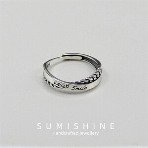 Könnte beinhalten: Verstellbarer Silberring mit der Aufschrift „keep Smile“ auf dem Band. Der Ring hat auf einer Seite ein geflochtenes Design und wird auf weißem Hintergrund präsentiert. Handgefertigter Schmuck.