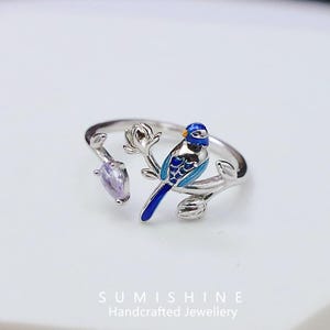 May include: A silver-toned ring featuring a blue and turquoise bird perched on a branch with a purple gemstone. The bird has intricate details, and the ring is set against a white background. The ring is handcrafted jewelry.