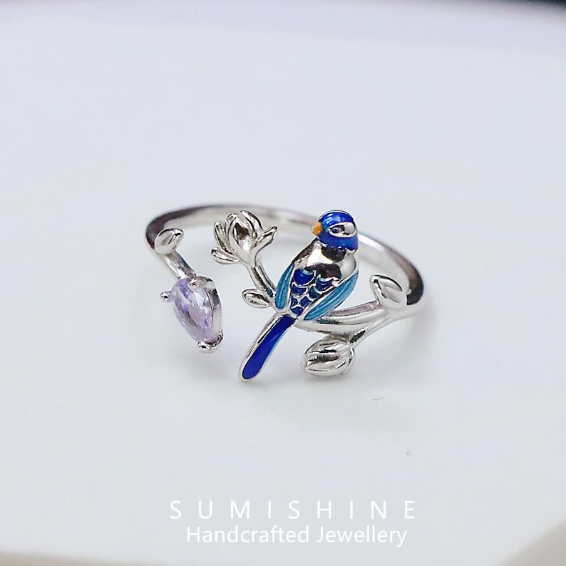 May include: A silver-toned ring featuring a blue and turquoise bird perched on a branch with a purple gemstone. The bird has intricate details, and the ring is set against a white background. The ring is handcrafted jewelry.