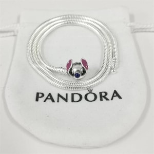 May include: A silver necklace with a charm depicting a cartoon character with pink ears, a blue nose, and black eyes. The necklace is coiled on a white pouch with the word "PANDORA" printed in black.
