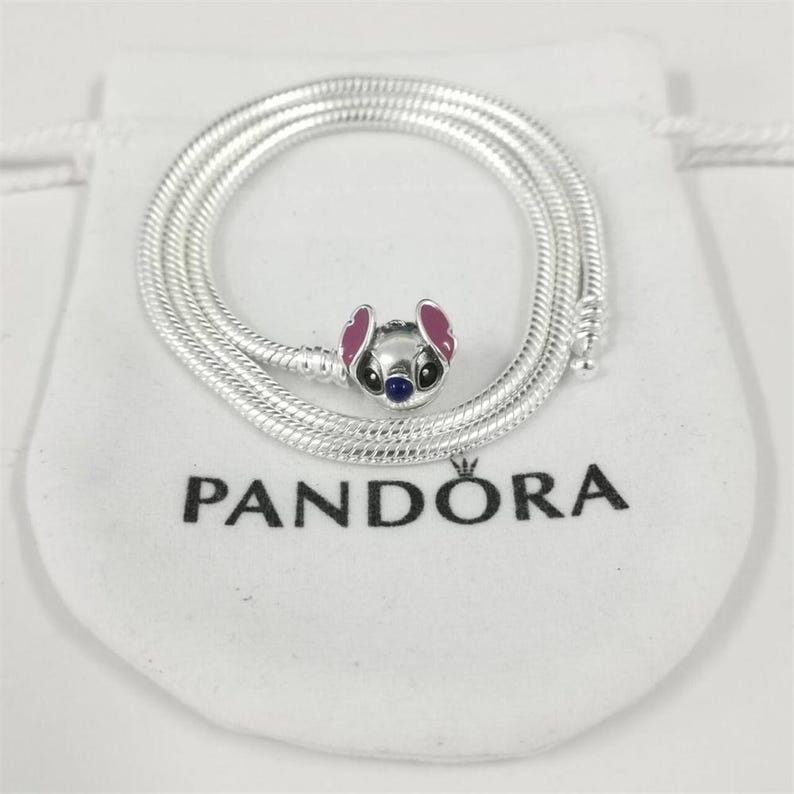 May include: A silver necklace with a charm depicting a cartoon character with pink ears, a blue nose, and black eyes. The necklace is coiled on a white pouch with the word "PANDORA" printed in black.