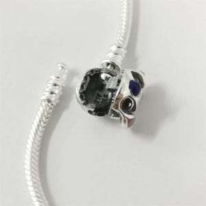 May include: A silver bracelet with a decorative cat-face charm. The charm has blue and pink details. The bracelet is unclasped, revealing the interlocking mechanism. The bracelet is made of silver.
