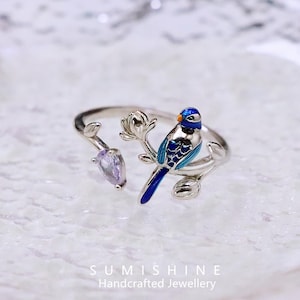 Könnte beinhalten: Silberfarbener Ring mit einem blauen Vogel auf einem Ast mit einem lila Edelstein. Der Vogel hat blaue und weiße Emaille-Details. Der Ring ist offen und verstellbar.