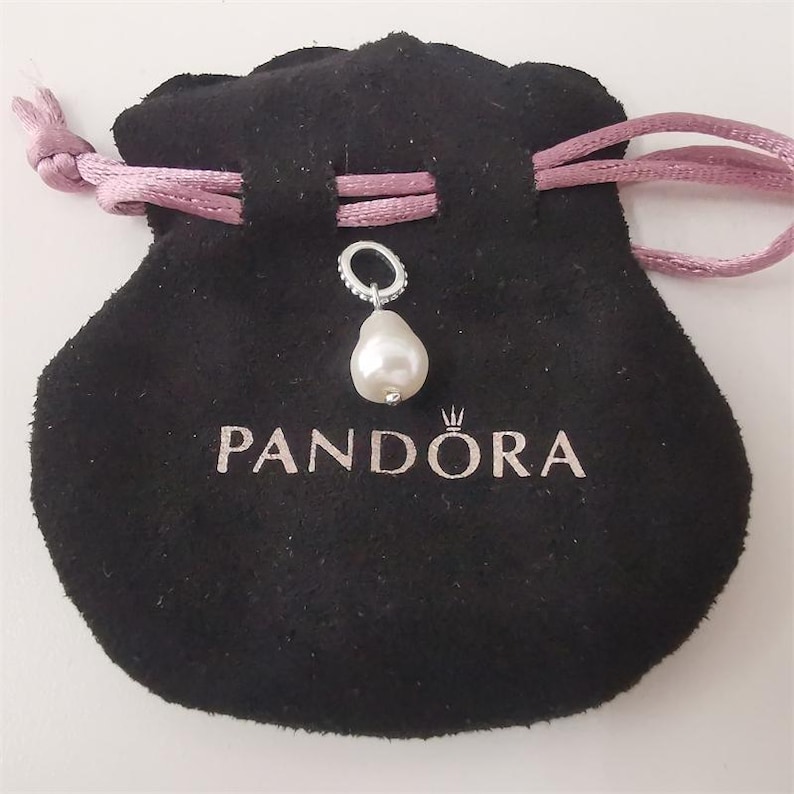 May include: A silver charm with a pear-shaped pearl pendant hangs above a black velvet pouch. The pouch has a pink drawstring and the word "PANDORA" printed in silver. The charm is attached to a silver ring.