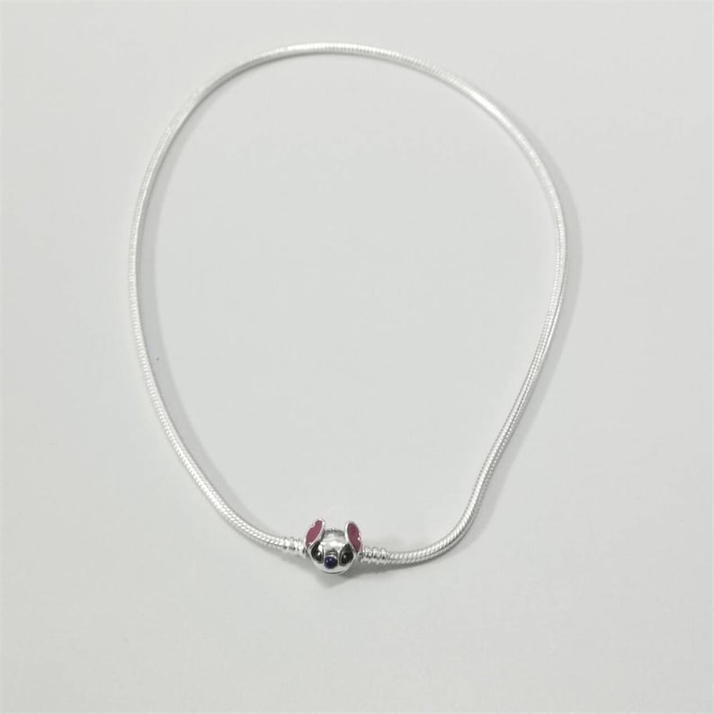 May include: A silver necklace with a charm depicting a cartoon character's face, with pink ears and a blue face. The necklace is made of a thin, flexible silver chain.