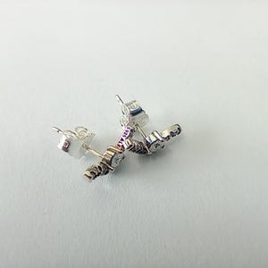May include: A pair of silver stud earrings with a decorative design. The earrings have a post backing and are set with clear gemstones. The metal is stamped with "925 ALE".