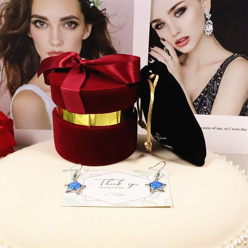 May include: A red velvet jewelry box with a satin bow, a black velvet pouch, and a pair of star-shaped earrings with blue centers. A card reads "Thank you" with a signature.