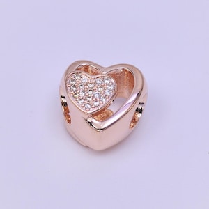 May include: A rose gold heart-shaped charm with a smaller heart filled with sparkling crystals. The charm has a polished finish and is designed with open spaces on the sides.